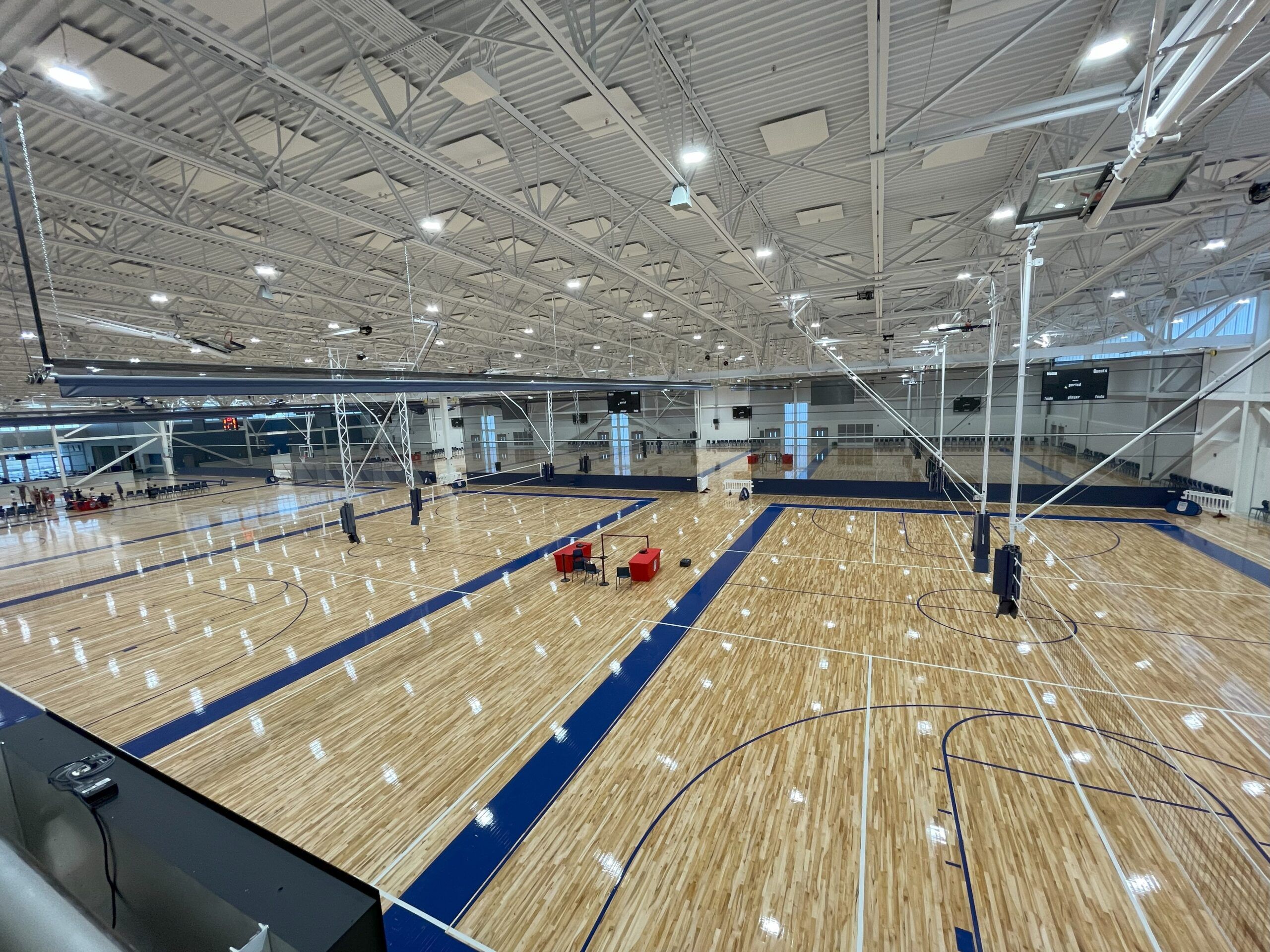 Multi-court hardwood gym floor installed by Sports Floors, Inc., with blue game lines, raised basketball goals, and divider curtains in a large athletic facility